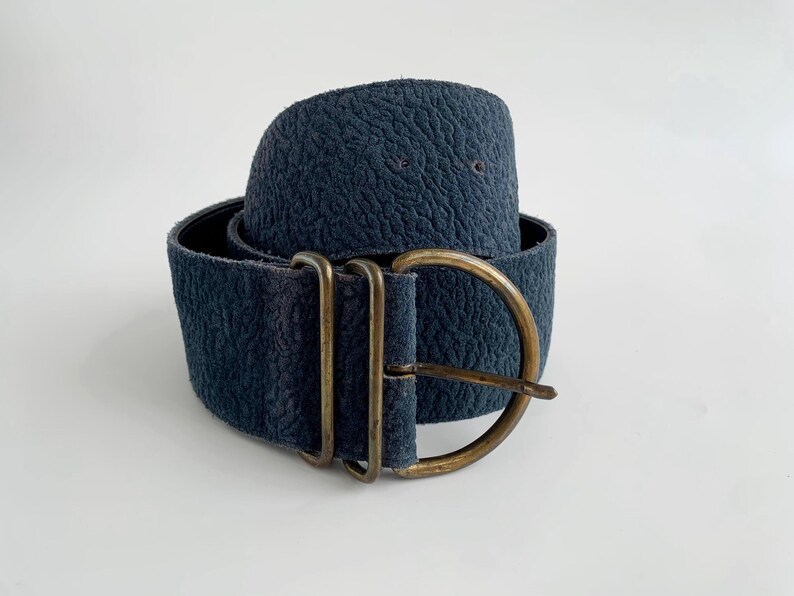 May include: A wide, navy blue belt with a textured surface. The belt features a large, antique brass buckle with a rectangular frame and a rounded buckle. The belt is approximately 7.6 cm wide.