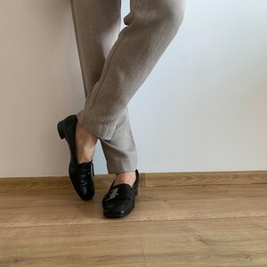 K&ouml;nnte beinhalten: Ein Paar schwarze Leder-Slipper mit silbernen Schnallen. Die Slipper werden zu einer beigefarbenen Hose getragen.