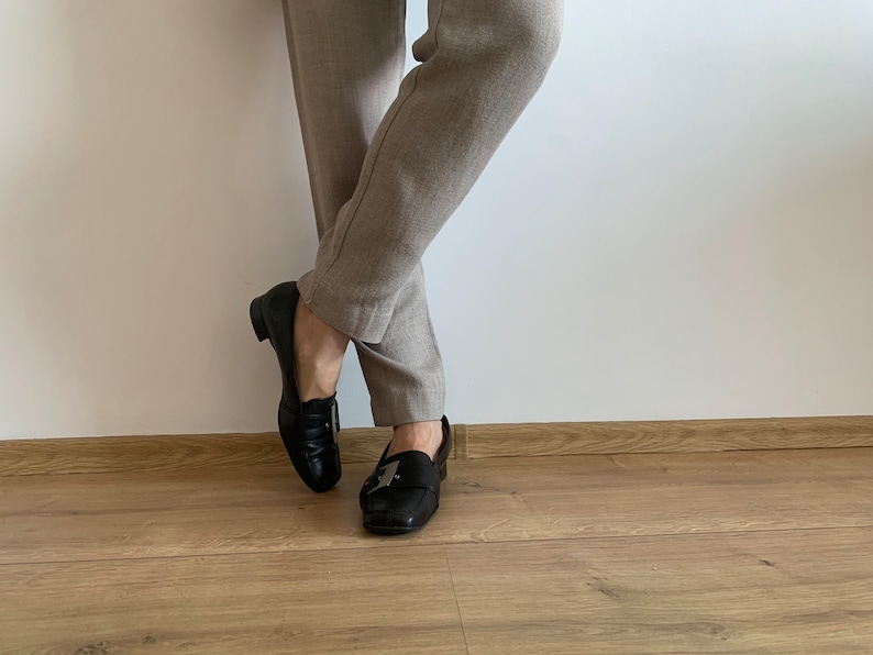 K&ouml;nnte beinhalten: Ein Paar schwarze Leder-Slipper mit silbernen Schnallen. Die Slipper werden zu einer beigefarbenen Hose getragen.