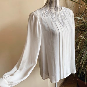 Puede incluir: Una blusa blanca de manga larga con cuello de encaje. La blusa tiene un frente plisado y est&aacute; hecha de una tela transparente.