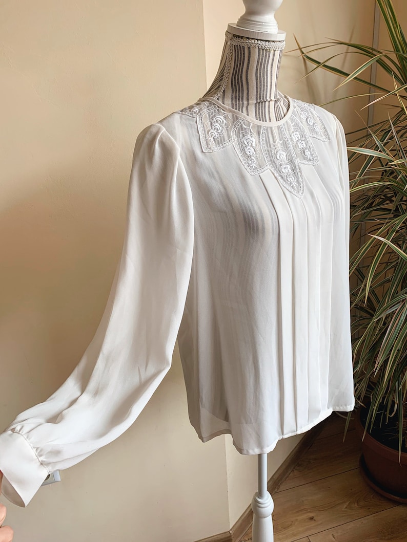 Puede incluir: Una blusa blanca de manga larga con cuello de encaje. La blusa tiene un frente plisado y est&aacute; hecha de una tela transparente.