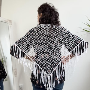 May include: A white, open-weave shawl with a diamond pattern and long fringe. The shawl is draped over a black floral print top and blue jeans. The shawl is a triangular shape.