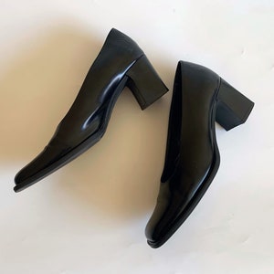 May include: A pair of black leather pumps with a square toe and a block heel. The heels are approximately 3 inches high. The shoes are glossy and appear unworn, set against a white background.