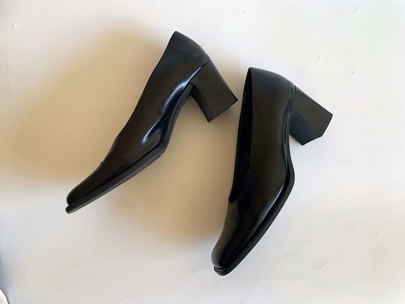 May include: A pair of black leather pumps with a square toe and a block heel. The heels are approximately 3 inches high. The shoes are glossy and appear unworn, set against a white background.