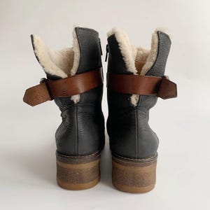 May include: A pair of black leather ankle boots featuring a cream-coloured faux fur lining, a brown leather strap with a metal buckle around the ankle, and a thick, textured brown rubber sole. The boots are shown from the back view.