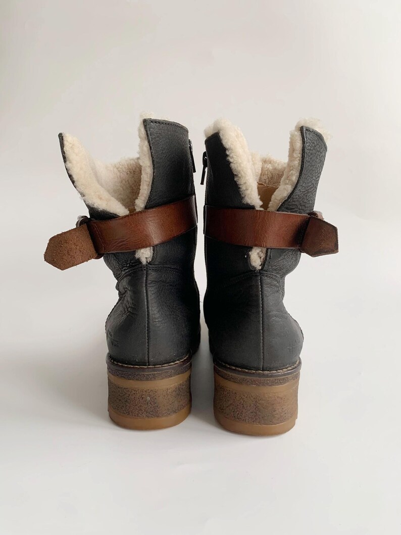 May include: A pair of black leather ankle boots featuring a cream-coloured faux fur lining, a brown leather strap with a metal buckle around the ankle, and a thick, textured brown rubber sole. The boots are shown from the back view.