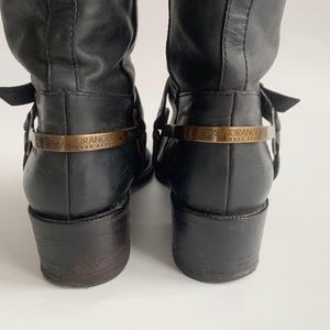 May include: A pair of black leather boots featuring a low block heel and a metallic gold-toned strap across the heel counter. The strap is engraved with the text BOSS ORANGE HUGO BOSS. The boots have a smooth finish and a structured design.