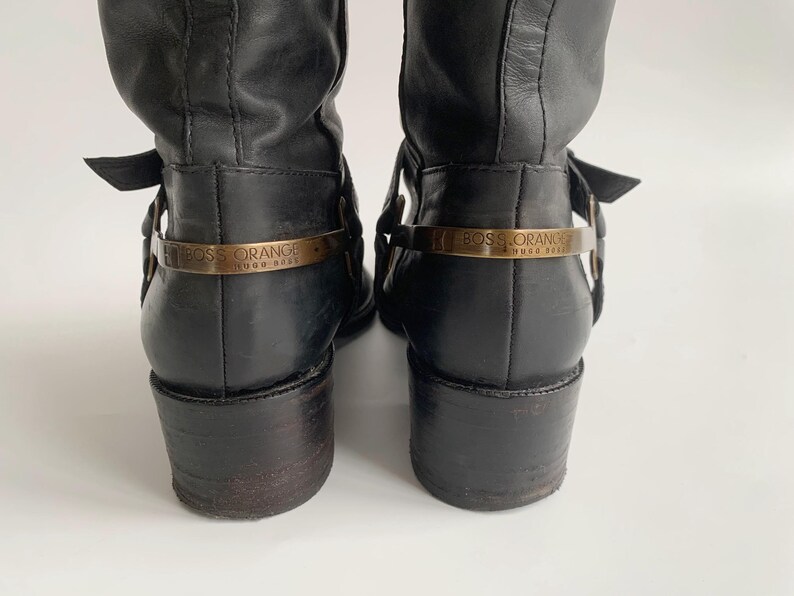 May include: A pair of black leather boots featuring a low block heel and a metallic gold-toned strap across the heel counter. The strap is engraved with the text BOSS ORANGE HUGO BOSS. The boots have a smooth finish and a structured design.