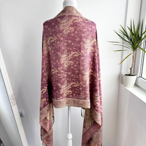 May include: A burgundy paisley patterned shawl draped over a white mannequin. The shawl features a detailed design in shades of beige and burgundy, with a fringed edge. The shawl is displayed in front of a white wall and a window.