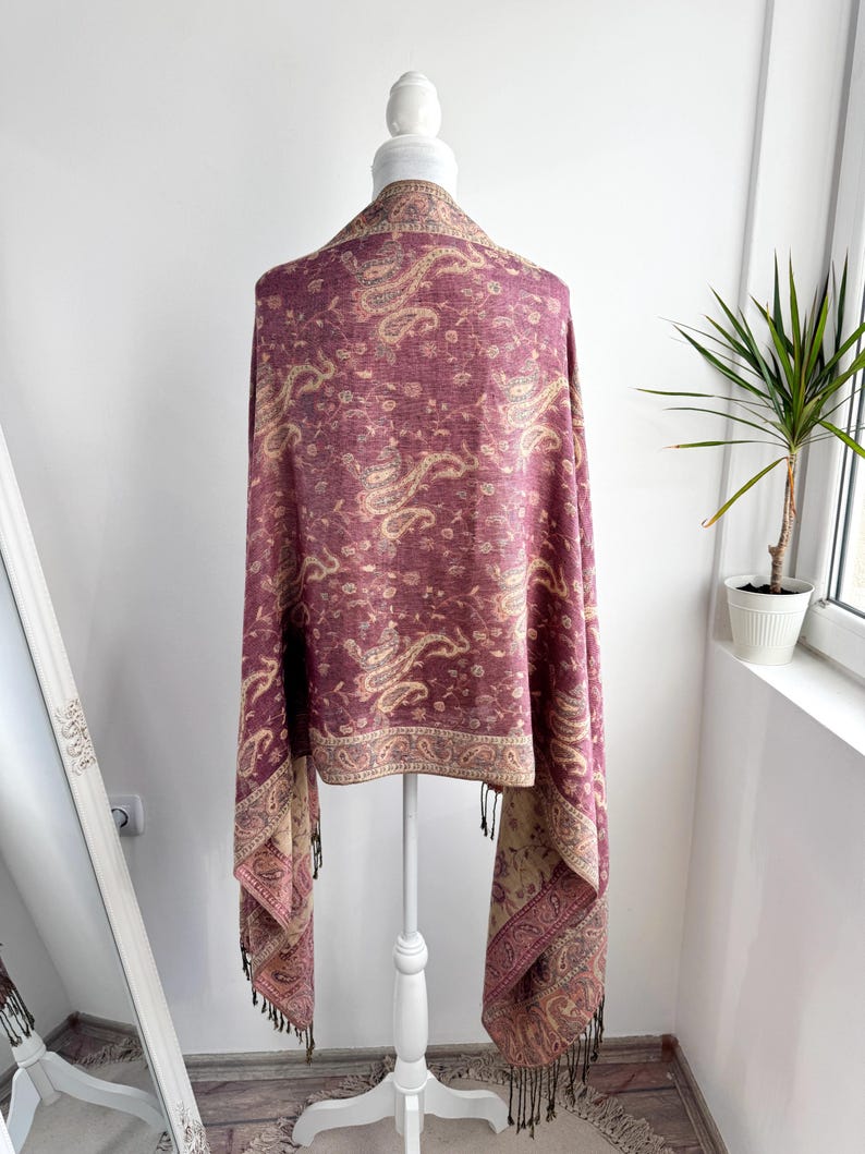 May include: A burgundy paisley patterned shawl draped over a white mannequin. The shawl features a detailed design in shades of beige and burgundy, with a fringed edge. The shawl is displayed in front of a white wall and a window.