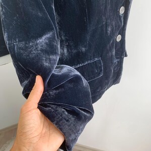 May include: A dark blue velvet blazer with silver buttons and a single pocket. The blazer is displayed on a white stand. The fabric has a soft, textured appearance.