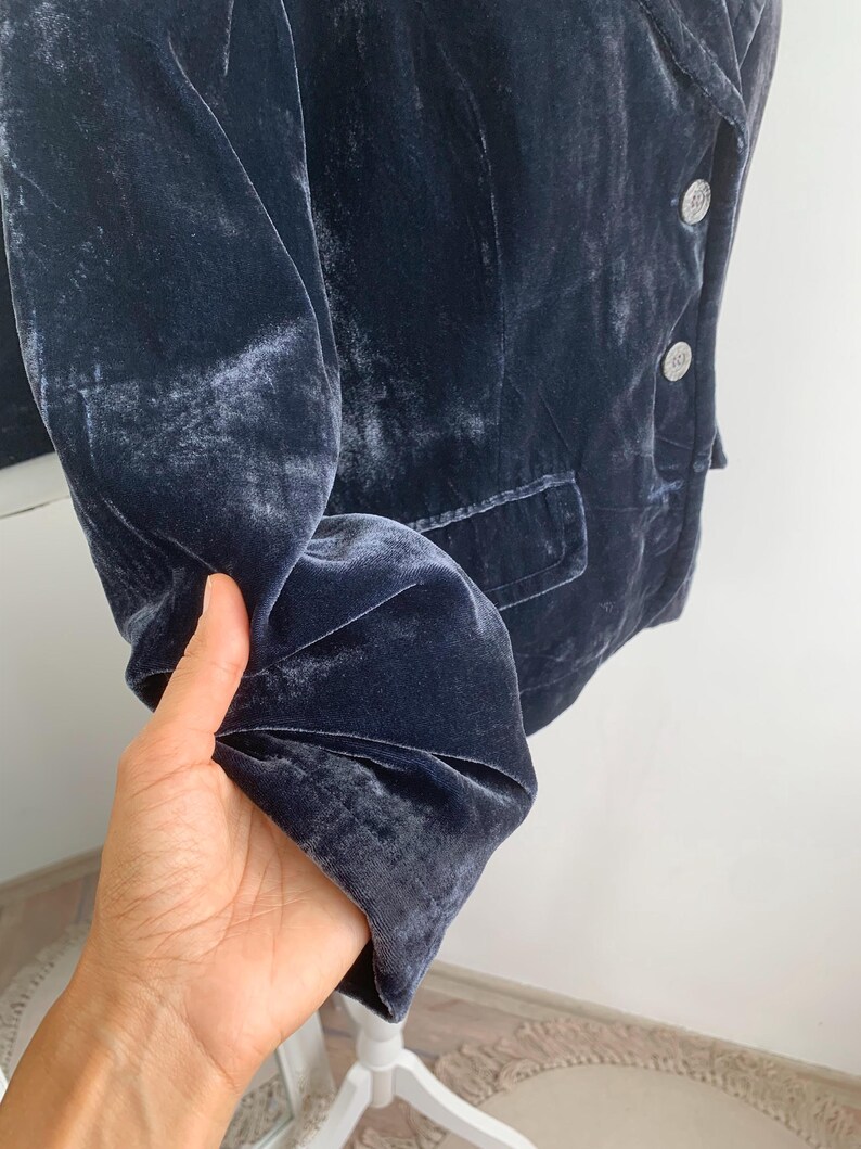 May include: A dark blue velvet blazer with silver buttons and a single pocket. The blazer is displayed on a white stand. The fabric has a soft, textured appearance.