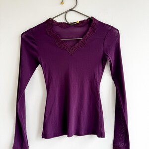 May include: A long-sleeved, deep purple shirt with a V-neck and delicate lace trim along the neckline and cuffs. The shirt is hanging on a black metal hanger against a white wall.