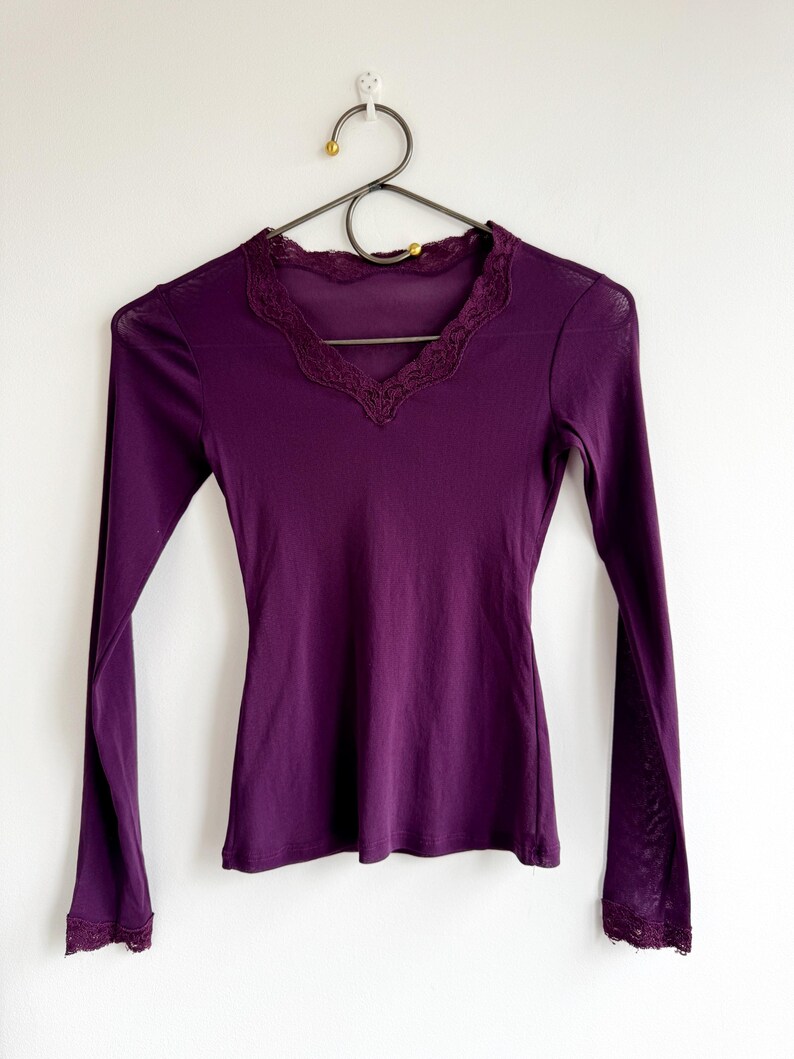 May include: A long-sleeved, deep purple shirt with a V-neck and delicate lace trim along the neckline and cuffs. The shirt is hanging on a black metal hanger against a white wall.