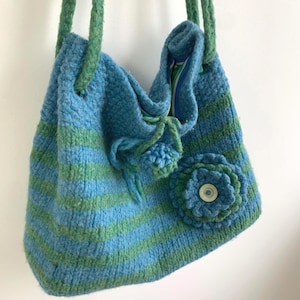 May include: A knitted bag in blue and green stripes, with green straps. The bag has a large flower decoration with a button in the centre and a small tassel. The interior lining is visible.