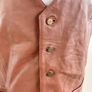 May include: A brown leather vest with a V-neck and four decorative buttons. The vest has a tailored fit and a pocket on the lower right side. The buttons are a gold color with a circular design.
