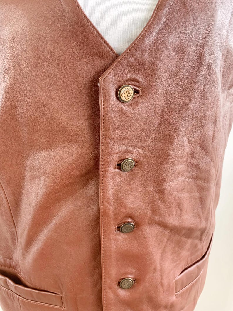 May include: A brown leather vest with a V-neck and four decorative buttons. The vest has a tailored fit and a pocket on the lower right side. The buttons are a gold color with a circular design.