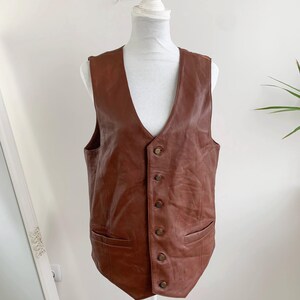 May include: A brown leather vest with a V-neck and button closure. The vest has two front pockets and is displayed on a mannequin. The leather has a slightly distressed appearance.