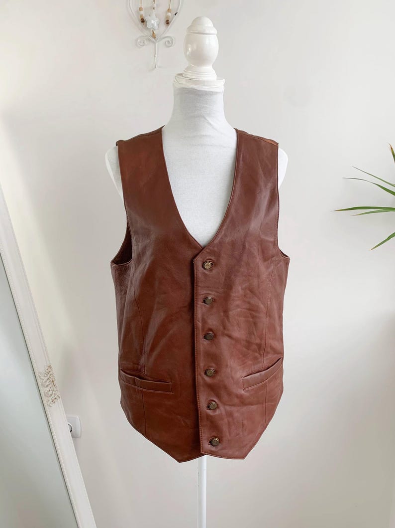 May include: A brown leather vest with a V-neck and button closure. The vest has two front pockets and is displayed on a mannequin. The leather has a slightly distressed appearance.