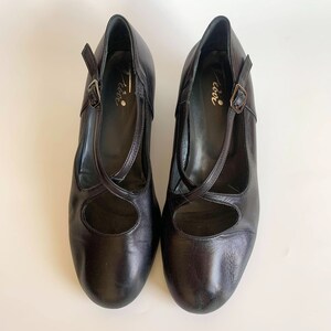Pu&ograve; includere: Un paio di scarpe in pelle nera con cinturino e fibbia. Le scarpe hanno punta tonda e tacco basso. Il nome del marchio "Coco" &egrave; scritto all'interno delle scarpe.