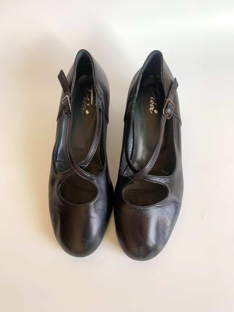 Pu&ograve; includere: Un paio di scarpe in pelle nera con cinturino e fibbia. Le scarpe hanno punta tonda e tacco basso. Il nome del marchio "Coco" &egrave; scritto all'interno delle scarpe.