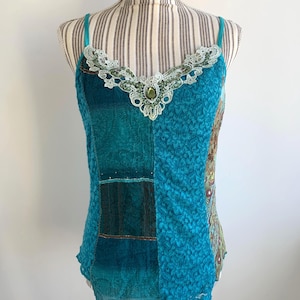 May include: A turquoise blue sleeveless top with a lace neckline and a green jewel accent. The top is made of a patchwork of different fabrics, including lace and velvet.
