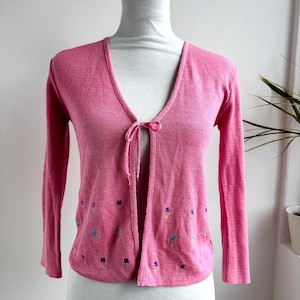 May include: A pink, long-sleeved cardigan with a V-neck and tie closure. The cardigan features small embroidered flowers in blue, purple, and green along the bottom hem. The garment is displayed on a white mannequin.