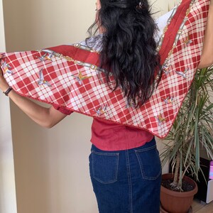 May include: A red and white plaid scarf with a pattern of birds and gold chains. The scarf is tied around the neck of a person wearing a red shirt and blue denim skirt.