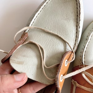 May include: A pair of white leather boat shoes with brown leather accents and white laces. The shoes have a casual, nautical style.