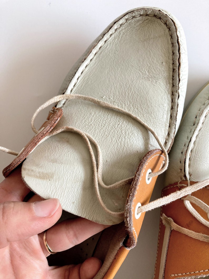 May include: A pair of white leather boat shoes with brown leather accents and white laces. The shoes have a casual, nautical style.