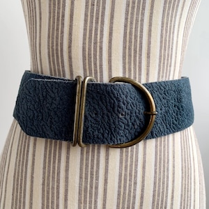 May include: A wide, navy blue belt with a textured surface and a large, antique brass-coloured buckle. The belt is displayed on a mannequin with a striped fabric pattern.