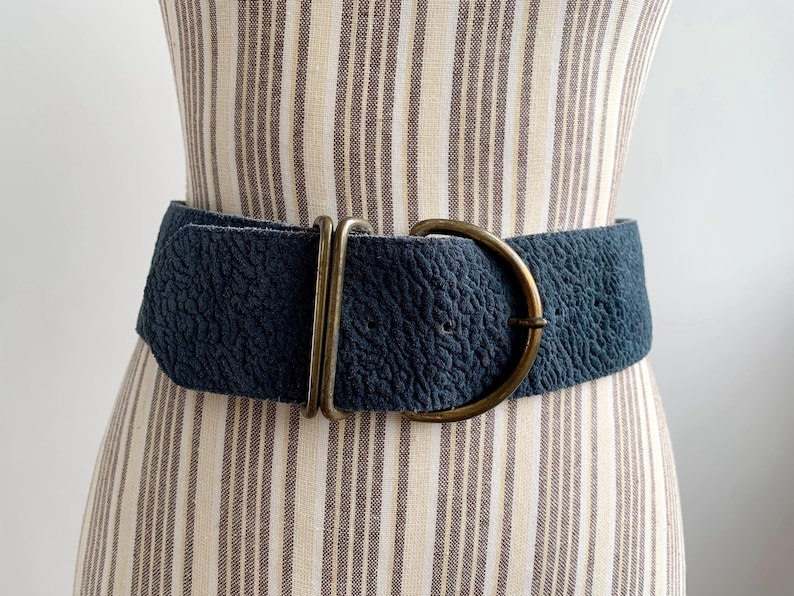 May include: A wide, navy blue belt with a textured surface and a large, antique brass-coloured buckle. The belt is displayed on a mannequin with a striped fabric pattern.