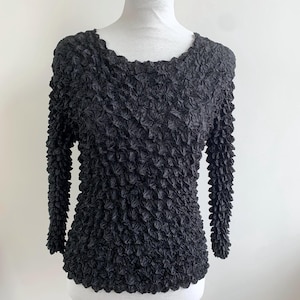 Puede incluir: Top negro de manga larga con un patrón texturizado en relieve. El top tiene un escote redondo y se muestra en un maniquí blanco. La tela tiene un diseño dimensional único, creando un efecto visualmente interesante.