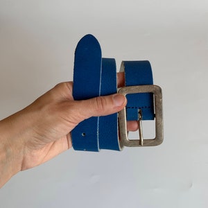 Pu&ograve; includere: Una cintura in pelle blu con una fibbia argentata. La cintura &egrave; piegata a met&agrave; ed &egrave; tenuta in una mano.