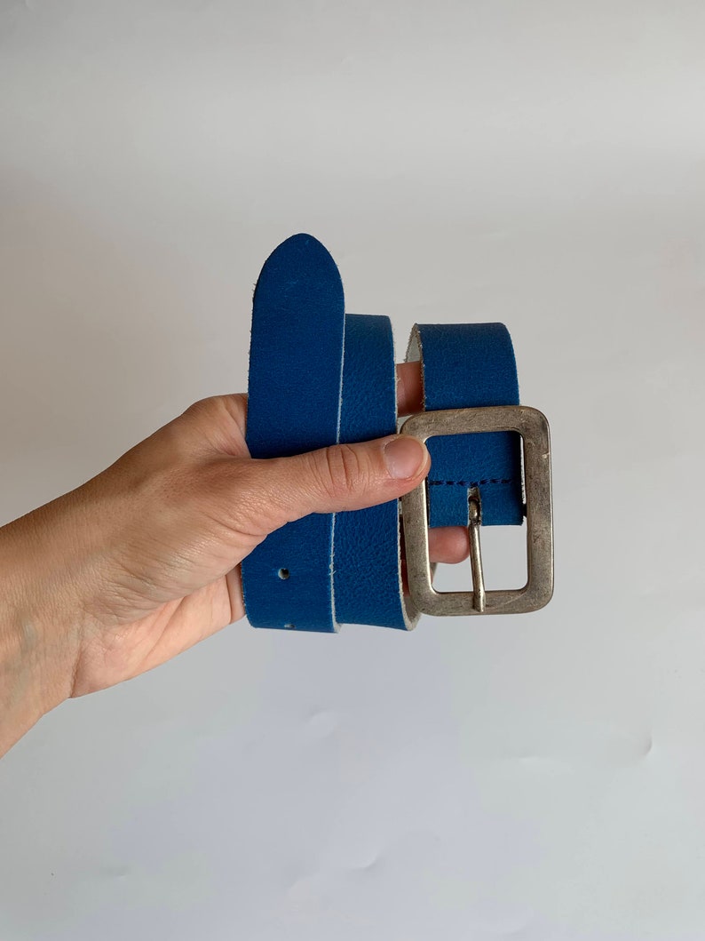 Pu&ograve; includere: Una cintura in pelle blu con una fibbia argentata. La cintura &egrave; piegata a met&agrave; ed &egrave; tenuta in una mano.