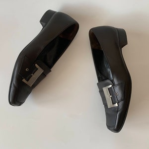 K&ouml;nnte beinhalten: Ein Paar schwarze Leder-Slipper mit silbernen Schnallenakzenten. Die Slipper haben eine spitze Zehenpartie und einen niedrigen Absatz.