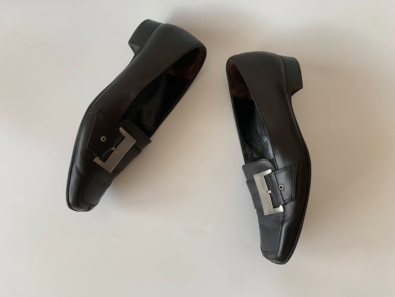 K&ouml;nnte beinhalten: Ein Paar schwarze Leder-Slipper mit silbernen Schnallenakzenten. Die Slipper haben eine spitze Zehenpartie und einen niedrigen Absatz.