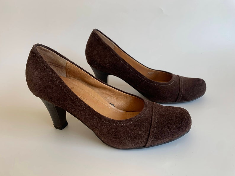 May include: A pair of brown suede heeled pumps with a closed toe and a rounded toe shape.