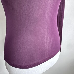 May include: A long-sleeved, sheer plum-colored top. The fabric is draped over a white mannequin, showcasing the garment's form-fitting design. The top has a rounded hemline and a subtle sheen, suggesting a lightweight material.