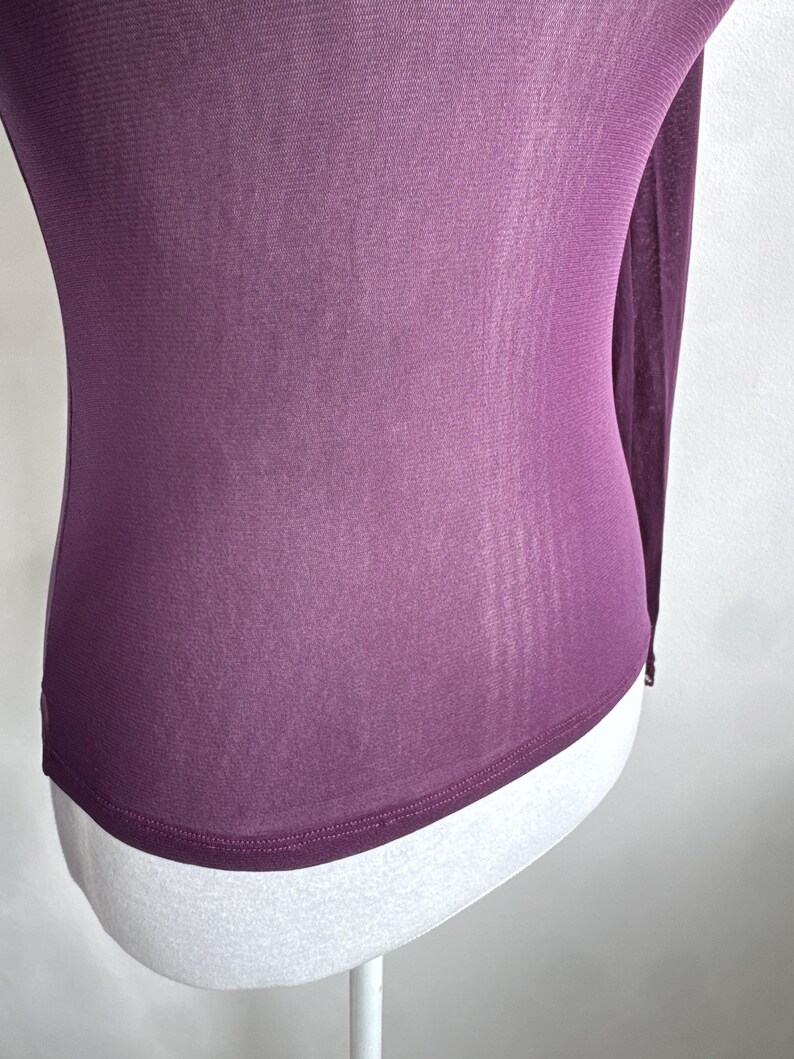 May include: A long-sleeved, sheer plum-colored top. The fabric is draped over a white mannequin, showcasing the garment's form-fitting design. The top has a rounded hemline and a subtle sheen, suggesting a lightweight material.