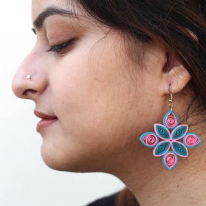May include: A pair of pink and teal blue paper quilled earrings in the shape of a flower. The earrings are dangling from the ear of a person.