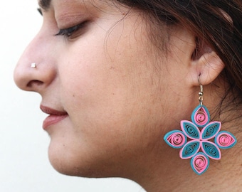 May include: A pair of pink and teal blue paper quilled earrings in the shape of a flower. The earrings are dangling from the ear of a person.