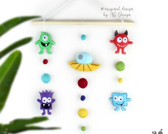 May include: A colorful felt mobile with a UFO, three different monsters, and colorful felt balls hanging from a wooden dowel. The mobile is suspended from a white string and a white hook. The text "original design by A6 Design" is printed on a white background.