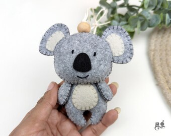Può includere: Un giocattolo di peluche koala in feltro grigio con occhi neri e pancia beige. Il koala tiene una piccola perla di legno nella sua mano.