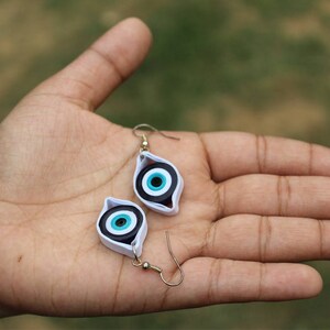 May include: A pair of earrings with a blue and white evil eye design. The earrings are made of paper and have a silver hook.