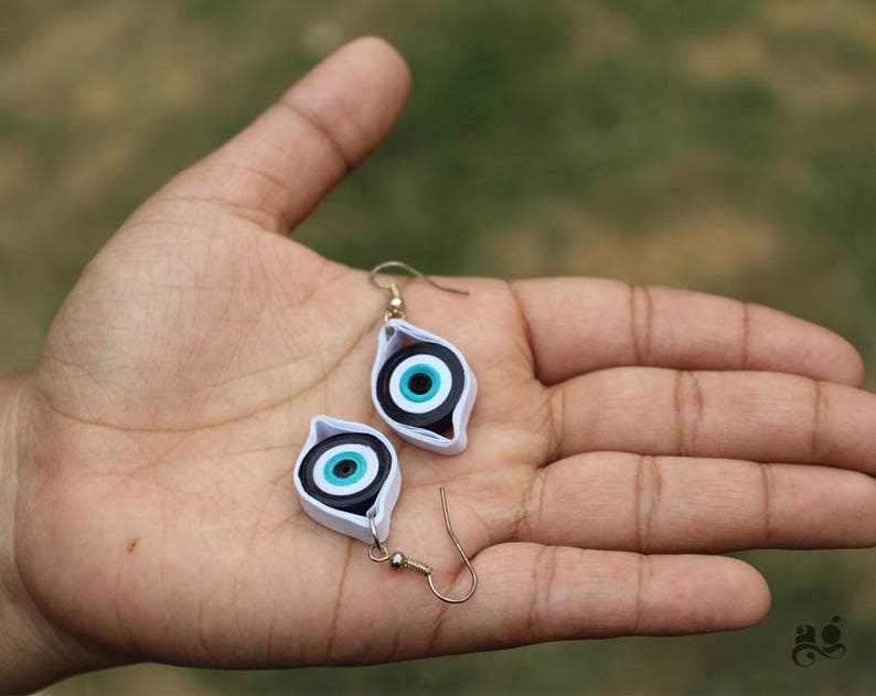 May include: A pair of earrings with a blue and white evil eye design. The earrings are made of paper and have a silver hook.
