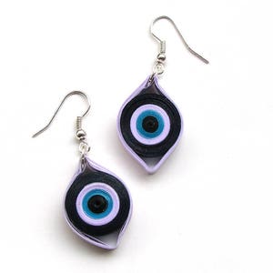 May include: A pair of earrings with a purple and black evil eye design. The earrings are made of paper and have silver hooks.