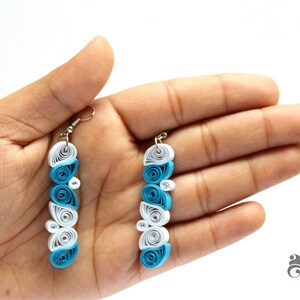 Può includere: Un paio di orecchini lunghi e pendenti realizzati con la tecnica del quilling. Gli orecchini sono bianchi e blu, con le sezioni blu nella parte inferiore degli orecchini. Gli orecchini sono appesi a ganci argentati.