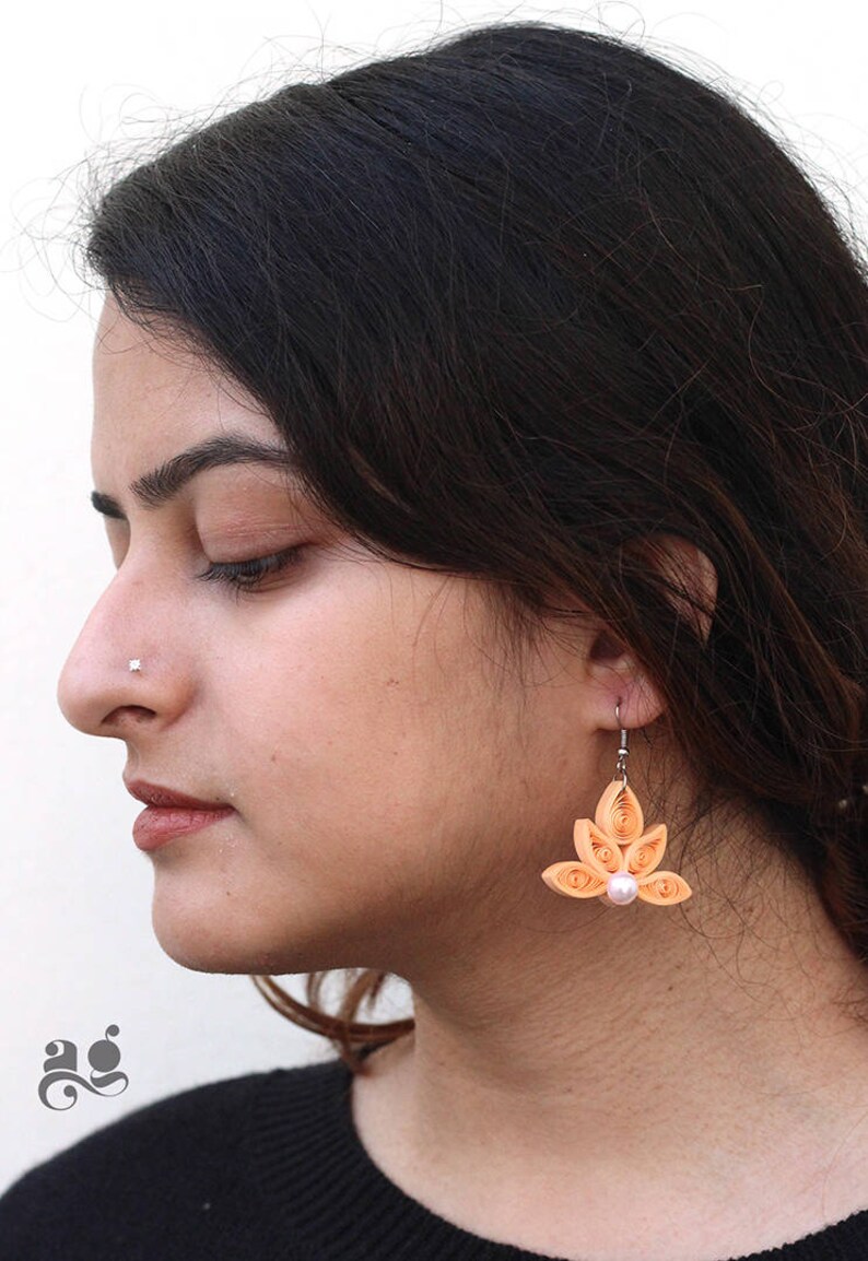 Pu&ograve; includere: Un paio di orecchini in quilling di carta color pesca con un accento di perla. Gli orecchini hanno la forma di un fiore con cinque petali.