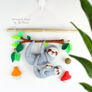 May include: A gray felt sloth with a baby sloth hanging on its back. The sloth is hanging from a wooden branch with green felt leaves. There are also two felt mushrooms, one orange and one red, hanging from the branch.